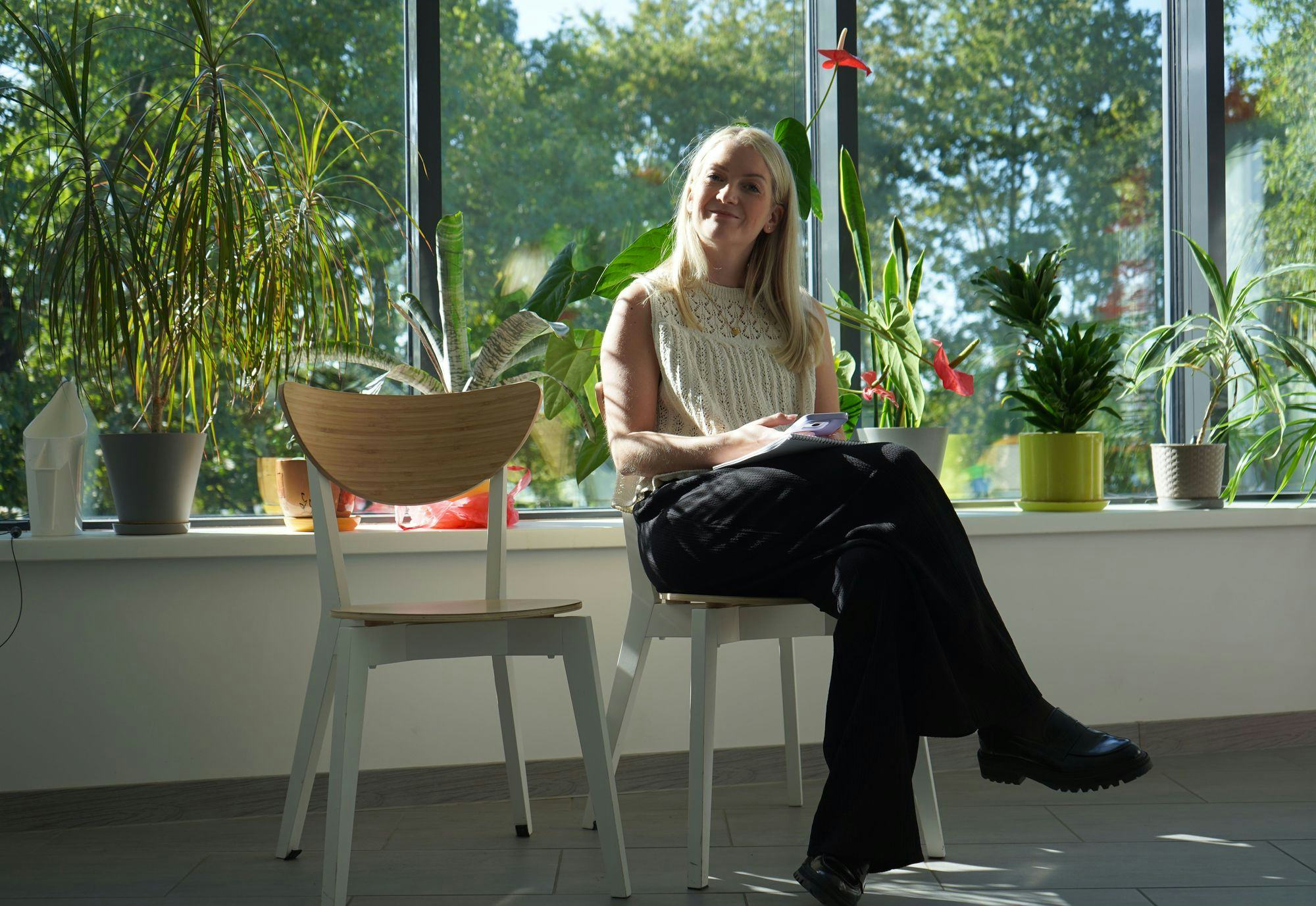 A woman sitting with her back to a window smiling, green plants all around
